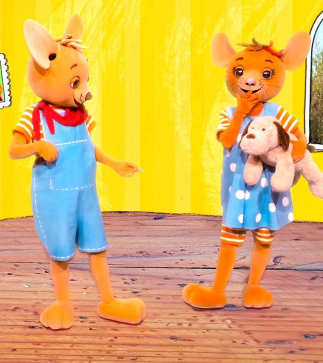 Jo (links) und Naya (rechts) stehen im gelben Raum. Naya hält einen Kuscheltier-Hund im Arm. Draußen am Fenster sind grüne Büsche zu erkennen.