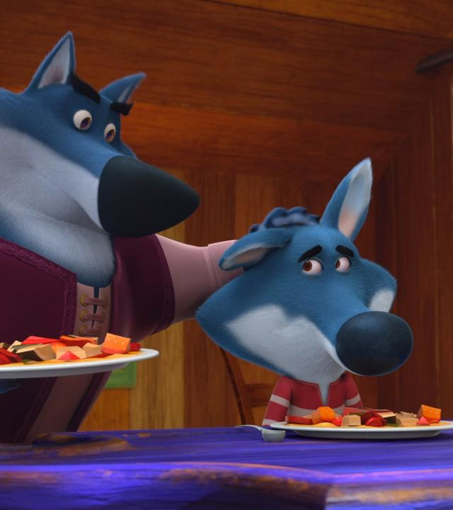 Der große böse Wolf streicht seinem Sohn Paulo über den Kopf. Sie haben Teller voller Gemüse vor sich.