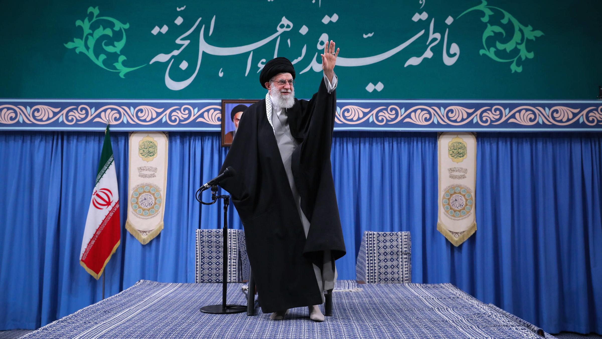 Ali Hosseini Khamenei steht auf einer Bühne, die mit Teppich ausgelegt ist. Er trägt eine schwarze Robe und winkt. Im Hintergrund steht die iranische Flagge.