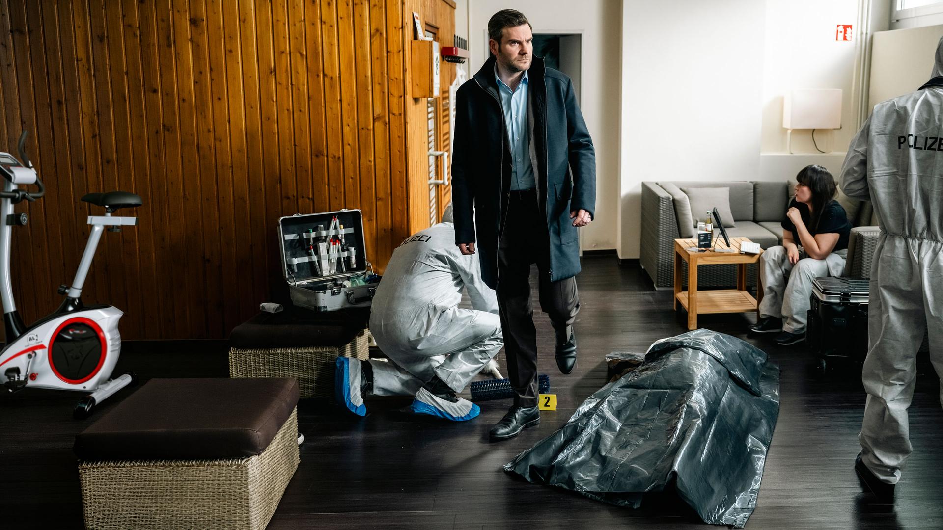 "SOKO Köln - Wer Kollegen hat, braucht keine Feinde": Jonas Fischer (Lukas Piloty) läuft in einem Wohnraum neben einer Leiche, die mit einer dunkelgrauen Plane abgedeckt wurde. Er sieht verwundert zur Seite. Im Hintergrund sitzt auf einem hellgrauen Sofa Dr. Pilar Westphal (Yulia Yáñez Schmidt), die vor sich auf ein Tablet sieht. Hinter Jonas kniet ein Mitarbeiter (Komparse) der Spurensicherung. Neben diesem steht ein aufgeklappter Koffer mit Arbeitsmaterialien. An der Seite steht ein weiterer Mitarbeiter (Komparse) der Spurensicherung.