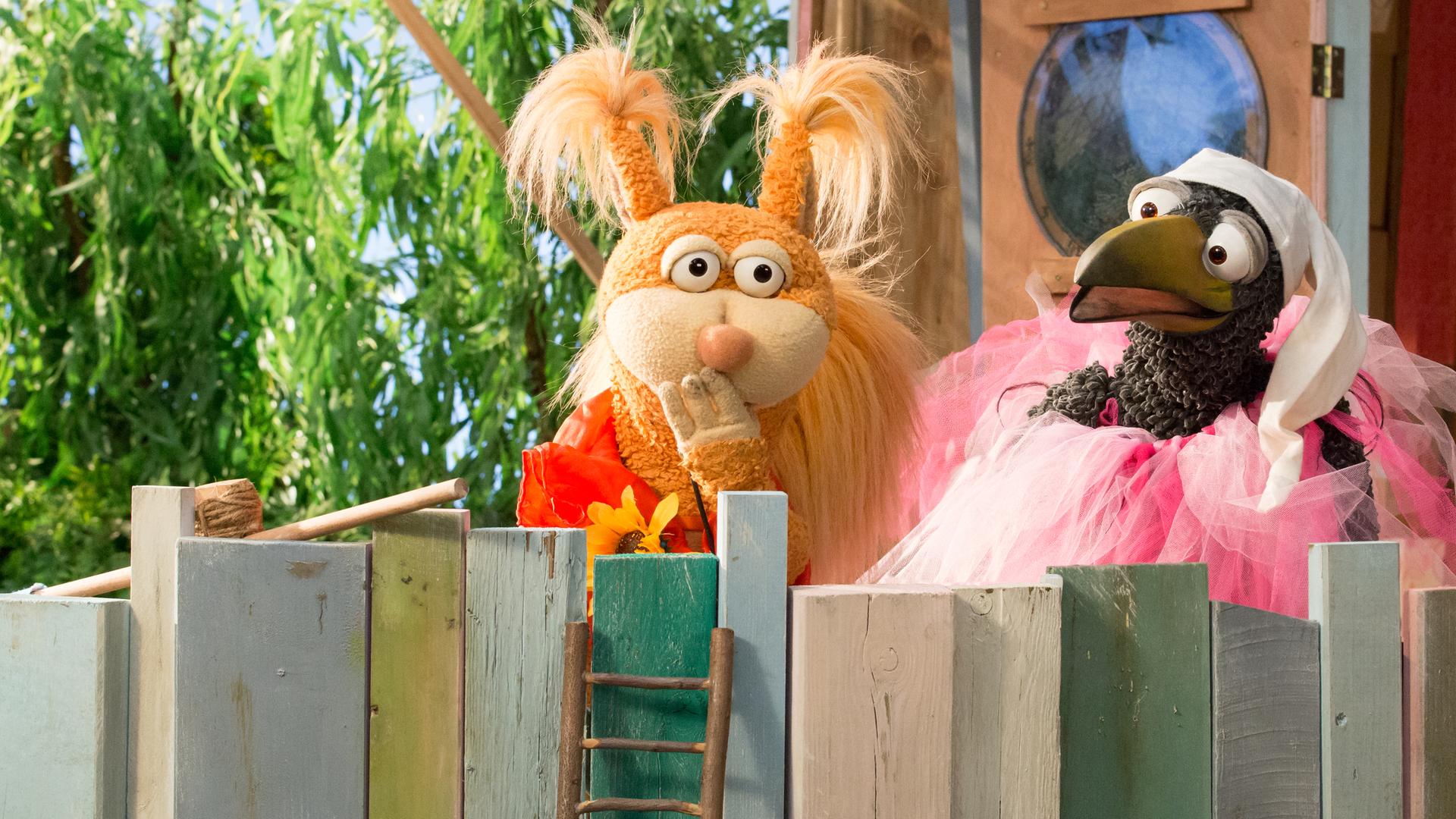 Rudi und Ella sind oben beim Baumhaus. Rudi trägt ein rosa Tütü und eine Schlafmütze. Ella hält die Hand vor den Mund, um sich ein Lachen zu verkneifen.