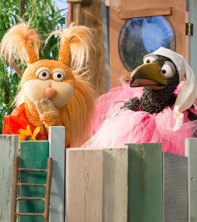 Rudi und Ella sind oben beim Baumhaus. Rudi trägt ein rosa Tütü und eine Schlafmütze. Ella hält die Hand vor den Mund, um sich ein Lachen zu verkneifen.