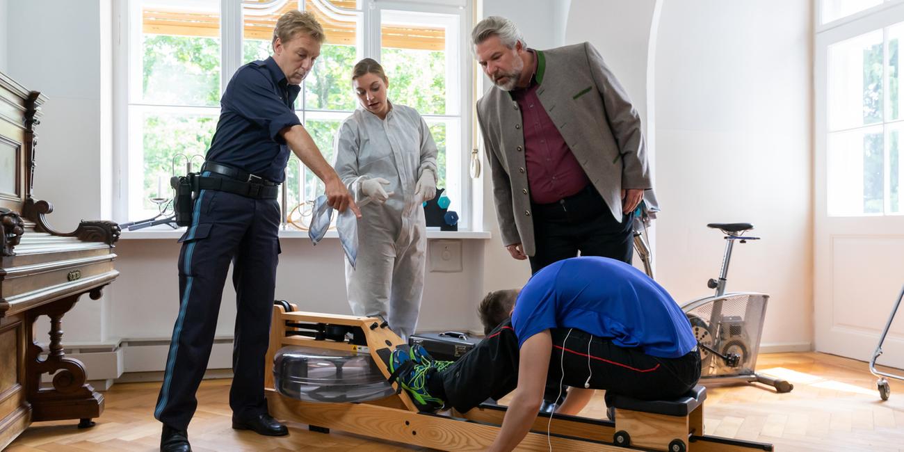 "Die Rosenheim-Cops - Wer rechnet schon mit Mord": Polizeihauptmeister Mohr (Max Müller), die Spurensicherin (Julia Angeli) und Kommissar Stadler (Dieter Fischer) stehen in einem Wohnraum um ein Trainingsgerät herum, auf dem das Opfer nach vorn gebeugt sitzt. Mohr zeigt auf das Smartphone, das neben der Leiche liegt.