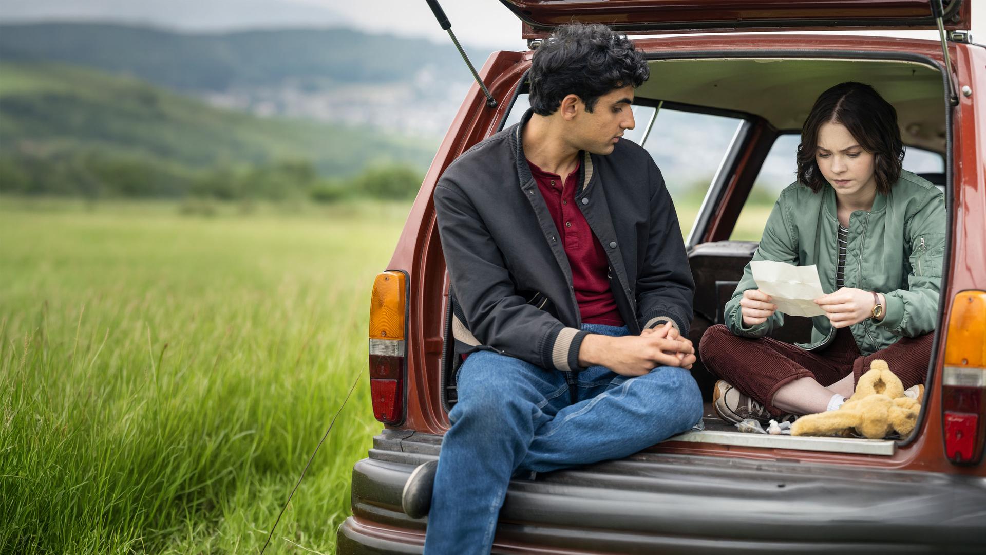 „A Good Girl's Guide to Murder – Wer war Andie Bell?“: Ravi (Zain Iqbal) und Pip (Emma Myers) sitzen im geöffneten Kofferraum eines roten Autos, das in einem Feld parkt. Pip studiert mit ernstem Blick ein weißes, aufgefaltetes Blatt, während Ravi sie von links daneben betrachtet. Vor Pip liegt zudem ein gelbes Kuscheltier.