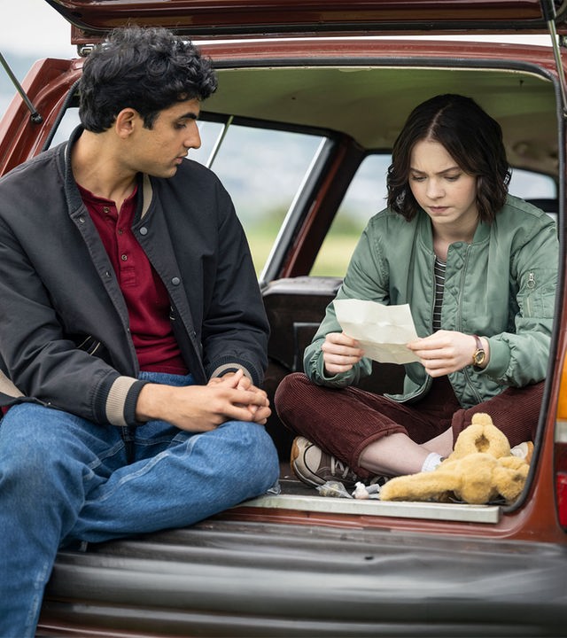 „A Good Girl's Guide to Murder – Wer war Andie Bell?“: Ravi (Zain Iqbal) und Pip (Emma Myers) sitzen im geöffneten Kofferraum eines roten Autos, das in einem Feld parkt. Pip studiert mit ernstem Blick ein weißes, aufgefaltetes Blatt, während Ravi sie von links daneben betrachtet. Vor Pip liegt zudem ein gelbes Kuscheltier.
