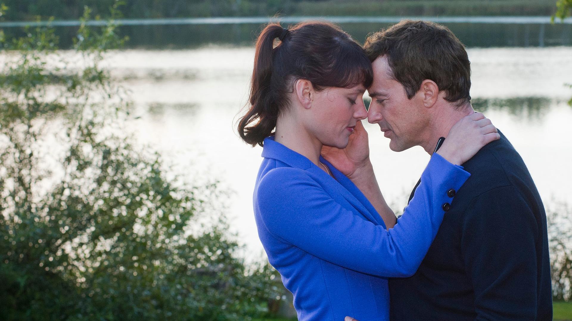 Pernilla (Maike Möller-Bornstein) und Jasper (Gedeon Burkhard) stehen sich umarmend auf einer Wiese vor einem See.