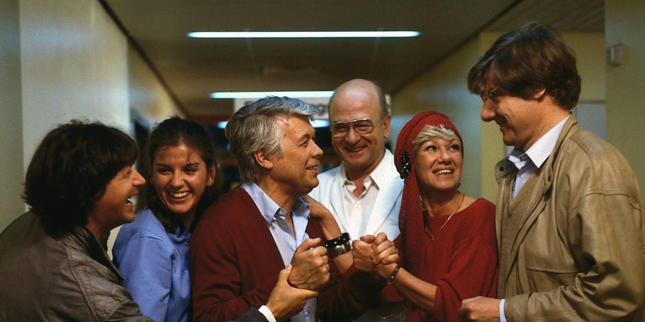 Eine Gruppe lächelnder Menschen schart sich um Werner. Dieser lächelt auch und alle scheinen sich über etwas zu freuen.