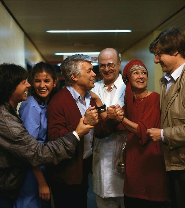 Eine Gruppe lächelnder Menschen schart sich um Werner. Dieser lächelt auch und alle scheinen sich über etwas zu freuen.