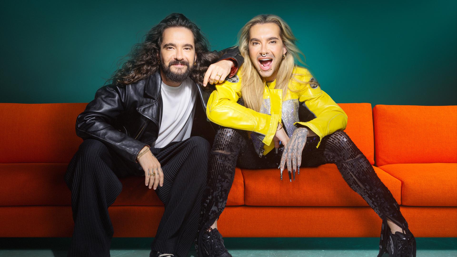 Tom und Bill Kaulitz sitzen nebeneinander auf einer roten Couch. Tom legt seinen linken Arm auf die rechte Schulter von Bill. Beide schauen in die Kamera.