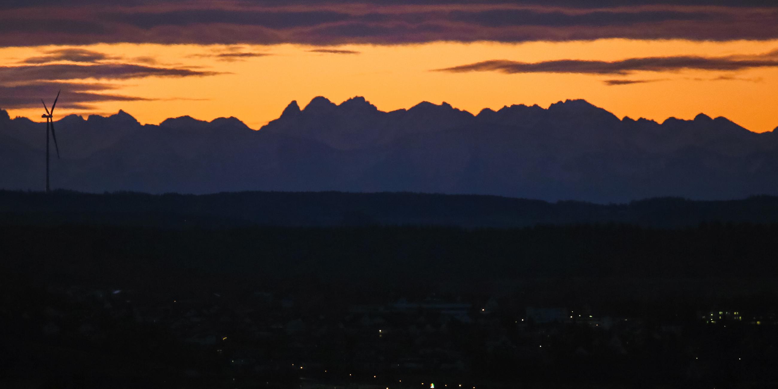 Während Oberschwaben noch im Dunkeln liegt, zeigen sich kurz vor Sonnenaufgang auf Grund der Föhnwetterlage hinter einem Windrad die über hundert Kilometer entfernten Alpen. 