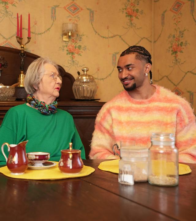 Eva (81) und Louis (26) sitzen an einem Tisch und schauen sich vertraut an. Im Hintergrund steht eine antike Anrichte.