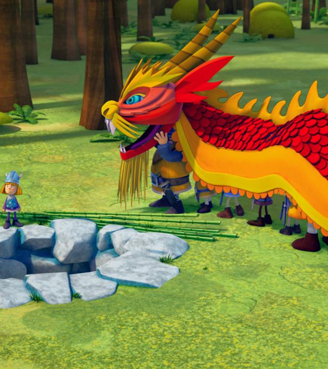 Die Wikinger haben sich als Drache verkleidet, um Ming und Qi zu helfen. Die beiden stehen links neben Wickie und sehen zum Drachenkostüm.