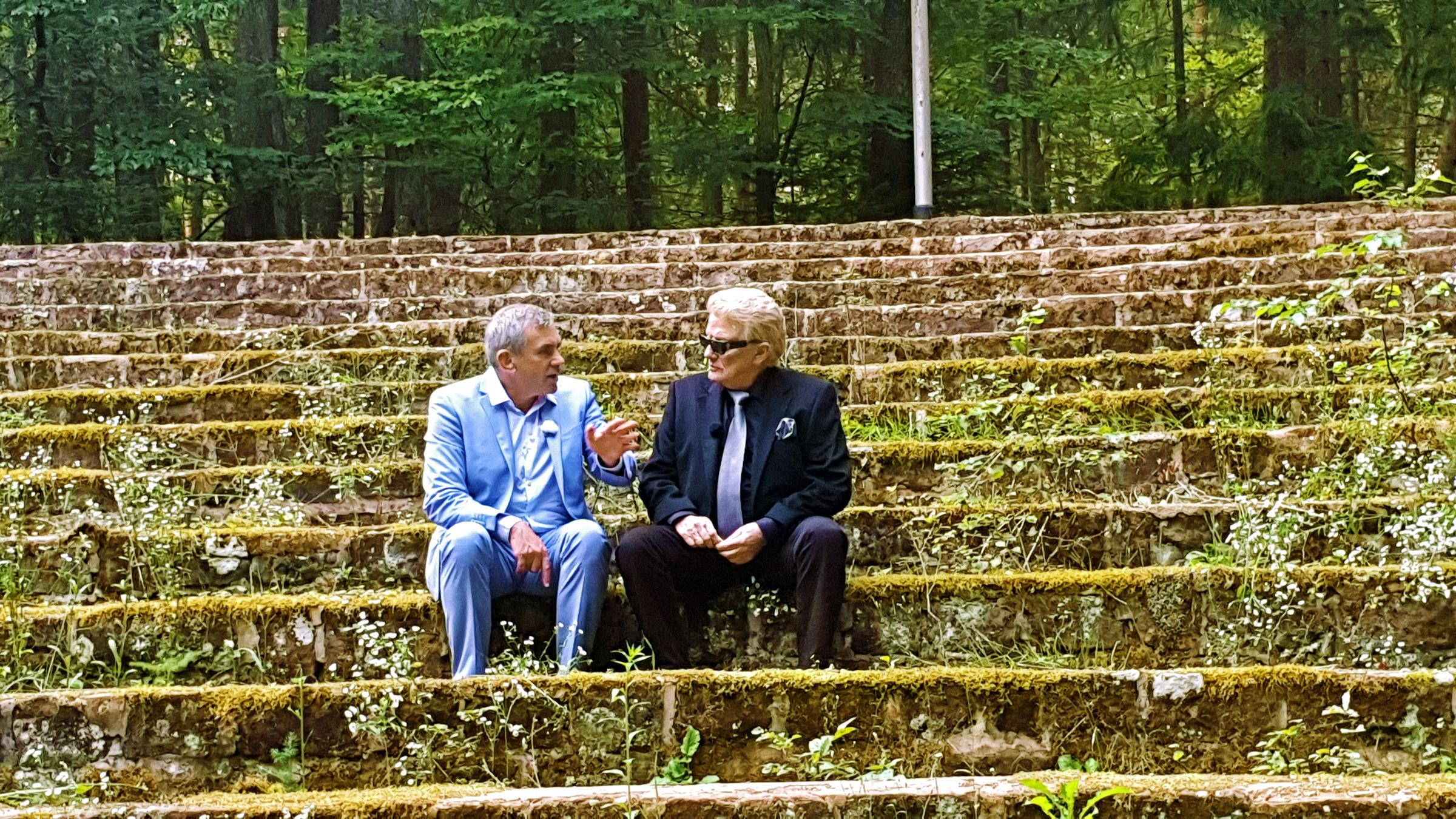 Wladimir Kamine und Heino sitzen auf einer steinernen, bemoosten Tribüne in einem Wald. Sie sind im Gespräch