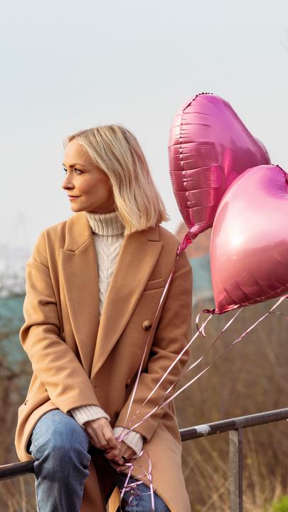 "Wie lange ist für immer?": Moderatorin Janin Ullmann sitzt auf einem Geländer, im Hintergrund ist der Hamburger Hafen zu sehen. Sie lässt den Blick in die Ferne schweifen. In den Händen hält sie die Fäden von vier Helium-Ballons in Herzform.