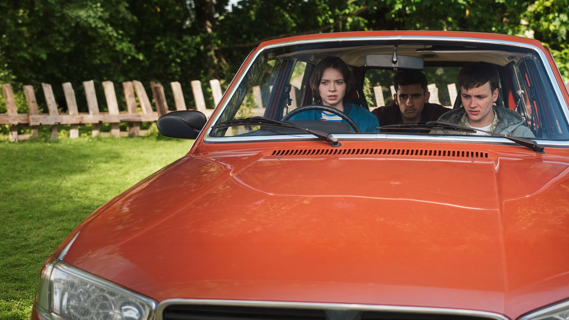 "A Good Girl’s Guide to Murder – Wie man einen Catfish angelt": Pip Fitz-Amobi (Emma Myers) sitzt am Steuer eines roten Autos, neben ihr sitzt Connor Reynolds (Jude Morgan-Collie), auf dem Rücksitz Ravi Singh (Zain Iqbal). Alle drei blicken angespannt nach vorn.
