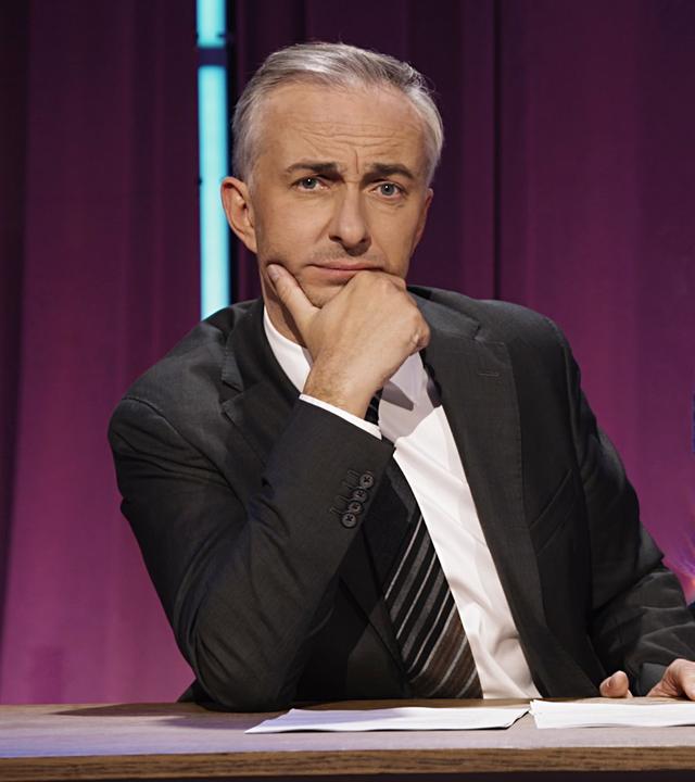 Jan Böhmermann sitzt am Schreibtisch. Rechts im Hintergrund sind die Silhouetten von drei Männern zu sehen.