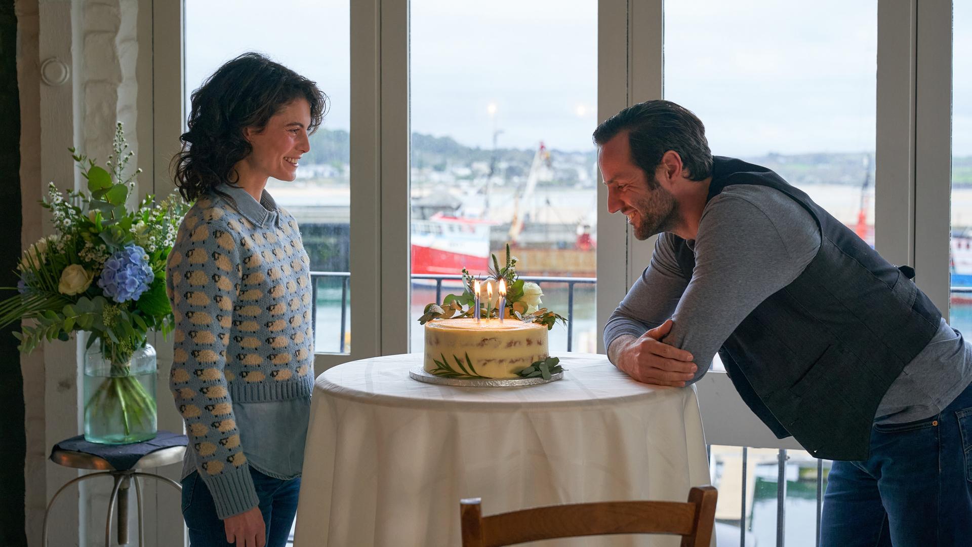 "Rosamunde Pilcher: Wie verhext": Grace (Jördis Richter) steht Sebastian (Stefan König) gegenüber, der sich vorbeugt, um die brennenden Kerzen auf einem Kuchen auszupusten. Im Hintergrund blickt man durch ein großes Fenster auf Schiffe im Hafen.