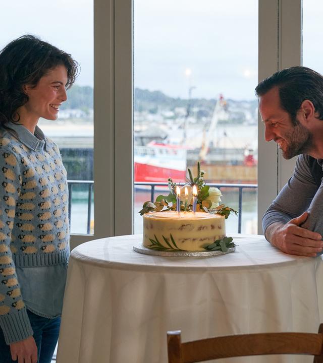 "Rosamunde Pilcher: Wie verhext": Grace (Jördis Richter) steht Sebastian (Stefan König) gegenüber, der sich vorbeugt, um die brennenden Kerzen auf einem Kuchen auszupusten. Im Hintergrund blickt man durch ein großes Fenster auf Schiffe im Hafen.