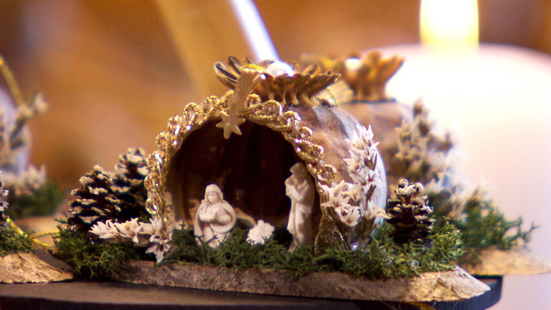 Das Bild zeigt eine kunstvoll gestaltete Krippe, die aus Mohnkapseln gefertigt ist. Die Krippe hat die Form eines kleinen, gewölbten Daches und ist mit goldfarbenen Verzierungen und einem kleinen Stern an der Spitze dekoriert. Vor der Krippe befindet sich eine Darstellung der heiligen Familie, mit Maria, Joseph und dem Jesuskind. Um die Krippe herum sind kleine, kunstvolle Details wie weiße Pflanzen und Zweige sowie Miniatur-Tannenzapfen angeordnet, die die Szenerie lebendig wirken lassen. Im Hintergrund sind weich beleuchtete Kerzen und unscharfe, warme Farben zu erkennen, die eine festliche und ruhige Atmosphäre schaffen. Diese Darstellung vermittelt den besinnlichen Charakter der Advents- und Weihnachtszeit in Österreich.