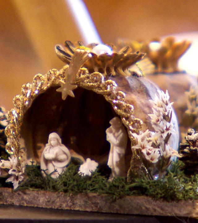 Das Bild zeigt eine kunstvoll gestaltete Krippe, die aus Mohnkapseln gefertigt ist. Die Krippe hat die Form eines kleinen, gewölbten Daches und ist mit goldfarbenen Verzierungen und einem kleinen Stern an der Spitze dekoriert. Vor der Krippe befindet sich eine Darstellung der heiligen Familie, mit Maria, Joseph und dem Jesuskind. Um die Krippe herum sind kleine, kunstvolle Details wie weiße Pflanzen und Zweige sowie Miniatur-Tannenzapfen angeordnet, die die Szenerie lebendig wirken lassen. Im Hintergrund sind weich beleuchtete Kerzen und unscharfe, warme Farben zu erkennen, die eine festliche und ruhige Atmosphäre schaffen. Diese Darstellung vermittelt den besinnlichen Charakter der Advents- und Weihnachtszeit in Österreich.