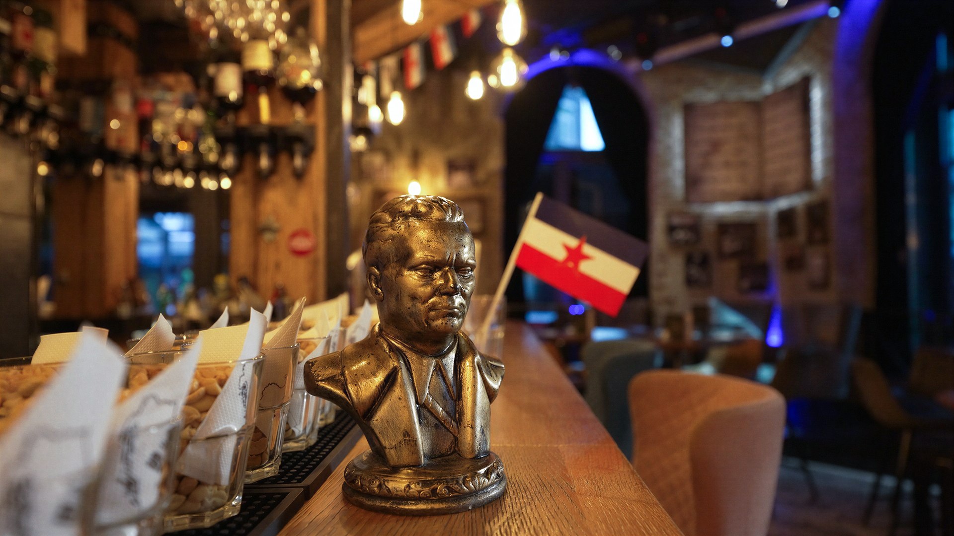 Das Bild zeigt die Inneneinrichtung eines Cafés namens „SRFJ“. Im Vordergrund befindet sich eine bronzeähnliche Büste eines männlichen Porträts, die auf der Bartheke steht. Die Büste zeigt einen Mann im Anzug mit einem ernsthaften Ausdruck. Hinter der Büste ist eine kleine Flagge mit einem roten Stern und horizontalen Streifen in Rot, Weiß und Blau zu sehen.  Im Hintergrund sind verschiedene Flaschen und Lichtquellen an der Decke sichtbar, die eine warme Atmosphäre schaffen. Der Raum hat eine rustikale Holzstruktur, und an der Wand hängen Bilder, die wahrscheinlich mit der Kultur des ehemaligen Jugoslawien in Verbindung stehen. Durch ein Fenster im Hintergrund ist Licht zu erkennen, das darauf hindeutet, dass es tagsüber ist.   Die Gesamtstimmung des Cafés scheint eine nostalgische und gemeinschaftliche Atmosphäre zu fördern, die durch die Einrichtung und die präsentierten Elemente zum Ausdruck kommt.