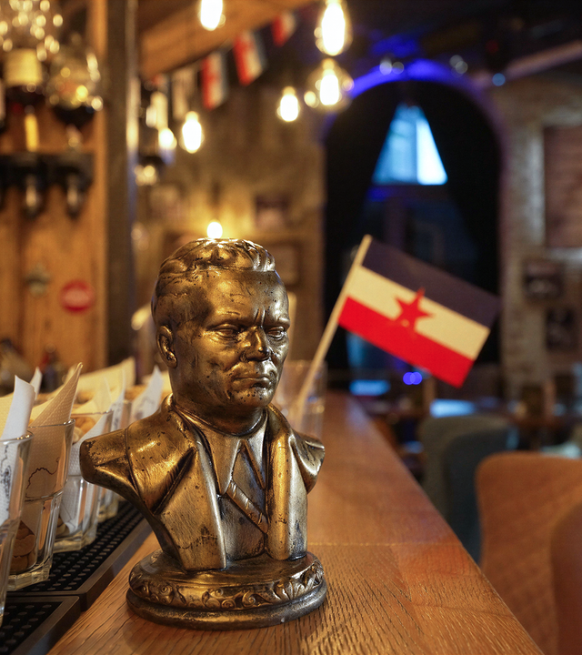 Das Bild zeigt die Inneneinrichtung eines Cafés namens „SRFJ“. Im Vordergrund befindet sich eine bronzeähnliche Büste eines männlichen Porträts, die auf der Bartheke steht. Die Büste zeigt einen Mann im Anzug mit einem ernsthaften Ausdruck. Hinter der Büste ist eine kleine Flagge mit einem roten Stern und horizontalen Streifen in Rot, Weiß und Blau zu sehen.  Im Hintergrund sind verschiedene Flaschen und Lichtquellen an der Decke sichtbar, die eine warme Atmosphäre schaffen. Der Raum hat eine rustikale Holzstruktur, und an der Wand hängen Bilder, die wahrscheinlich mit der Kultur des ehemaligen Jugoslawien in Verbindung stehen. Durch ein Fenster im Hintergrund ist Licht zu erkennen, das darauf hindeutet, dass es tagsüber ist.   Die Gesamtstimmung des Cafés scheint eine nostalgische und gemeinschaftliche Atmosphäre zu fördern, die durch die Einrichtung und die präsentierten Elemente zum Ausdruck kommt.