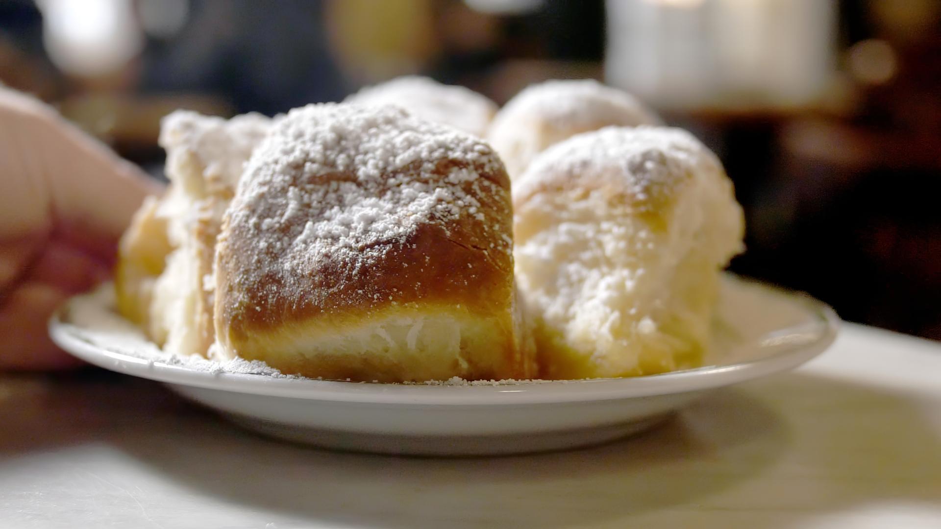 Sechs goldbraune Brötchen auf einem weißen Teller, jedes mit Puderzucker bestäubt, in einem kreisförmigen Muster angeordnet. Der Teller ruht auf einer weißen Tischdecke, und im Hintergrund ist eine verschwommene Hand zu erkennen.