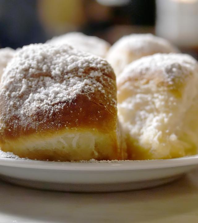 Sechs goldbraune Brötchen auf einem weißen Teller, jedes mit Puderzucker bestäubt, in einem kreisförmigen Muster angeordnet. Der Teller ruht auf einer weißen Tischdecke, und im Hintergrund ist eine verschwommene Hand zu erkennen.