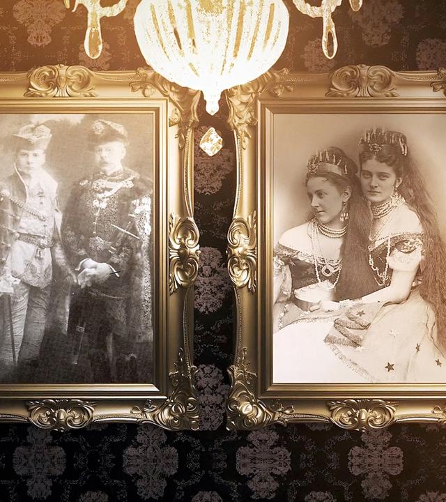 Das Bild zeigt eine ruhige Inneneinrichtung mit zwei gerahmten Fotografien, die an einer Wand hängen. Das linke Foto zeigt einen Mann und eine Frau in formeller Kleidung, wobei der Mann einen Gehstock hält und die Frau eine Tiara trägt. Das rechte Foto zeigt eine Frau in einem langen Kleid und einen Mann im Anzug, beide blicken direkt in die Kamera.