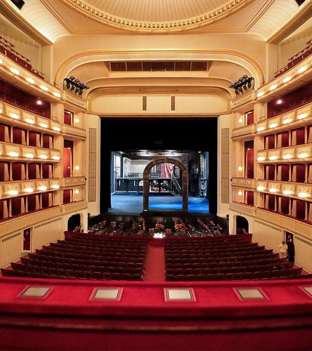 Das Bild zeigt den Innenraum der Wiener Staatsoper. Im Vordergrund sind rote gepolsterte Sitzplätze sichtbar, die in einem leicht geschwungenen Muster angeordnet sind. Über den Sitzplätzen befinden sich Balkone, die mit goldenen Verzierungen und rot-beige gestreiften Vorhängen dekoriert sind. An den Wänden sind mehrere Wandleuchten angebracht, die ein warmes Licht abgeben.  In der Mitte des Bildes ist die Bühne zu sehen, die dunkel und leer ist. Im Hintergrund des Bühnenszenarios sind technische Elemente und ein Teil des Bühnenbildes sichtbar, das eine architektonische Struktur zeigt.   Die gesamte Atmosphäre des Raumes vermittelt ein Gefühl von Eleganz und Tradition. Darüber hinaus kann man die Bühne in ihrer Vorbereitungsphase erkennen, da einige Techniker im Hintergrund zu sehen sind. Der Raum wirkt groß und offen, gekennzeichnet durch hohe Decken und aufwendig gestaltete Elemente.
