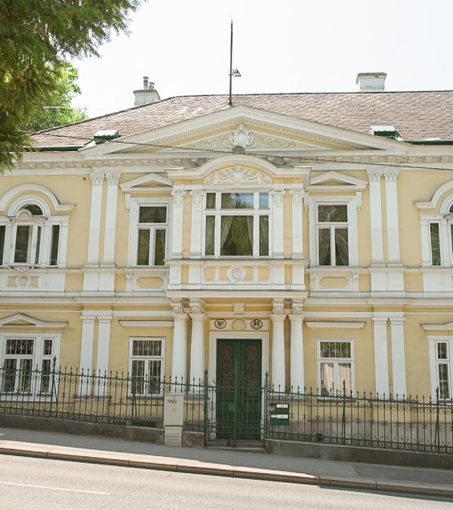 Das Bild zeigt die Fassade einer eleganten Villa in Hietzing, einem Stadtteil von Wien. Die Villa hat eine gelbe Außenwand mit weißen, kunstvollen Verzierungen, die den klassischen Stil des 19. Jahrhunderts repräsentieren.   In der Mitte der Fassade befindet sich der Eingang, der durch eine grüne Tür markiert ist. Über der Tür sind zwei große Fenster, die symmetrisch angeordnet sind. Weitere Fenster sind entlang der Fassade verteilt, einige mit Rundbögen.   Die Villa ist von einem niedrigen, grünen Zaun umgeben, der sich entlang der Straße erstreckt. Vor der Villa stehen einige hohe Bäume, die einen Teil der Sicht auf das Gebäude verdecken. Der Himmel ist klar und der Bereich insgesamt wirkt ruhig und wohlhabend.   In der EXIF-Beschreibung wird erwähnt, dass die Villa eine bedeutende historische Rolle spielt, da hier Kaiser Franz Joseph seine Geliebte besuchte.