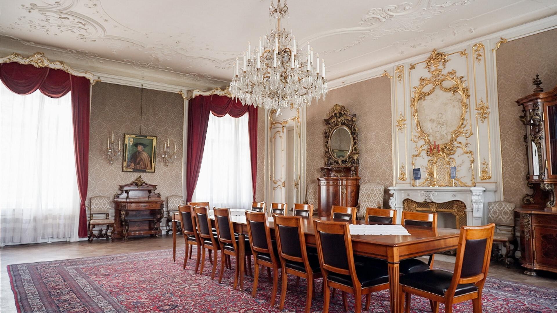 Das Bild zeigt einen eleganten, opulent gestalteten Raum im Augartenpalais. Der Raum ist groß und hat hohe Decken mit aufwendigen Verzierungen, die filigrane Stuckarbeiten aufweisen. An der Decke hängt ein großer, glitzernder Kronleuchter.  Die Wände sind mit einem zarten Muster in einem hellen Farbton dekoriert, und in einer der Wände ist ein großes, gerahmtes Porträt eines Mannes zu sehen. Flankiert von zwei Wandlampen ist dieser Bereich besonders hervorgehoben.   Vor dem Porträt befindet sich eine Holzkommode. Der Boden des Raumes ist mit einem persischen Teppich in warmen Rottönen ausgelegt. In der Mitte des Raumes steht ein großer, rechteckiger Tisch, umgeben von mehreren Holzstühlen mit schwarzen Sitzflächen.   Die Fenster sind mit schweren, roten Vorhängen dekoriert, die den Raum zusätzlich einladend wirken lassen. Auf der gegenüberliegenden Seite gibt es einen Kamin mit einem weiß verzierten Mantel und eine große, elegant geschwungene Holzvitrine, die antike Gegenstände beherbergt. Das gesamte Ambiente strahlt einen historischen und luxuriösen Charakter aus.