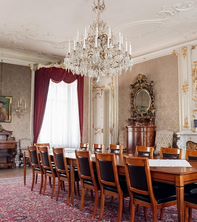 Das Bild zeigt einen eleganten, opulent gestalteten Raum im Augartenpalais. Der Raum ist groß und hat hohe Decken mit aufwendigen Verzierungen, die filigrane Stuckarbeiten aufweisen. An der Decke hängt ein großer, glitzernder Kronleuchter.  Die Wände sind mit einem zarten Muster in einem hellen Farbton dekoriert, und in einer der Wände ist ein großes, gerahmtes Porträt eines Mannes zu sehen. Flankiert von zwei Wandlampen ist dieser Bereich besonders hervorgehoben.   Vor dem Porträt befindet sich eine Holzkommode. Der Boden des Raumes ist mit einem persischen Teppich in warmen Rottönen ausgelegt. In der Mitte des Raumes steht ein großer, rechteckiger Tisch, umgeben von mehreren Holzstühlen mit schwarzen Sitzflächen.   Die Fenster sind mit schweren, roten Vorhängen dekoriert, die den Raum zusätzlich einladend wirken lassen. Auf der gegenüberliegenden Seite gibt es einen Kamin mit einem weiß verzierten Mantel und eine große, elegant geschwungene Holzvitrine, die antike Gegenstände beherbergt. Das gesamte Ambiente strahlt einen historischen und luxuriösen Charakter aus.