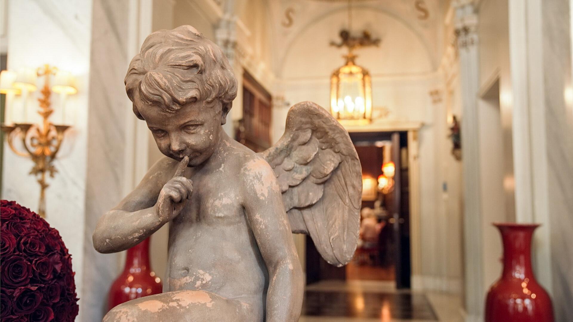 Das Bild zeigt eine innenarchitektonisch gestaltete Lobby eines eleganten Hotels, vermutlich das Hotel Sacher in Wien. Im Vordergrund steht eine Statue eines Engels, der leicht nachdenklich wirkt und mit einem Finger auf den Lippen den Ausdruck von Diskretion symbolisiert. Die Statue ist alt und hat eine matte, leicht abblätternde Oberfläche.   Im Hintergrund sind Wände mit hellen, möglicherweise marmorierten Oberflächen sichtbar, und eine elegante Lampe hängt von der Decke. Links vom Engel befinden sich rote Rosen, die in einem tiefen, runden Behälter arrangiert sind. Rechts stehen rote Vasen, die sich harmonisch ins Gesamtbild einfügen. Im Hintergrund sind verschwommene Umrisse von Tischen und Stühlen zu erkennen, was auf eine gestaltete Essens- oder Lounge-Atmosphäre hinweist. Die Lichtverhältnisse sind warm und laden zu einem entspannten Aufenthalt ein.