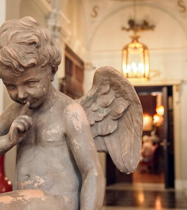 Das Bild zeigt eine innenarchitektonisch gestaltete Lobby eines eleganten Hotels, vermutlich das Hotel Sacher in Wien. Im Vordergrund steht eine Statue eines Engels, der leicht nachdenklich wirkt und mit einem Finger auf den Lippen den Ausdruck von Diskretion symbolisiert. Die Statue ist alt und hat eine matte, leicht abblätternde Oberfläche.   Im Hintergrund sind Wände mit hellen, möglicherweise marmorierten Oberflächen sichtbar, und eine elegante Lampe hängt von der Decke. Links vom Engel befinden sich rote Rosen, die in einem tiefen, runden Behälter arrangiert sind. Rechts stehen rote Vasen, die sich harmonisch ins Gesamtbild einfügen. Im Hintergrund sind verschwommene Umrisse von Tischen und Stühlen zu erkennen, was auf eine gestaltete Essens- oder Lounge-Atmosphäre hinweist. Die Lichtverhältnisse sind warm und laden zu einem entspannten Aufenthalt ein.