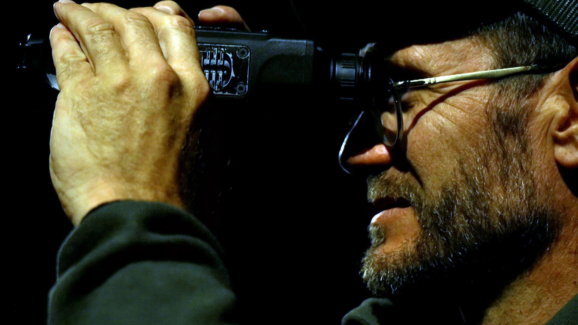 Nahaufnahme eines Mannes im Profil, der nachts durch ein Fernglas schaut. Er trägt eine Brille und eine Mütze, und seine Hand hält das Fernglas fest, während er konzentriert in die Dunkelheit blickt.