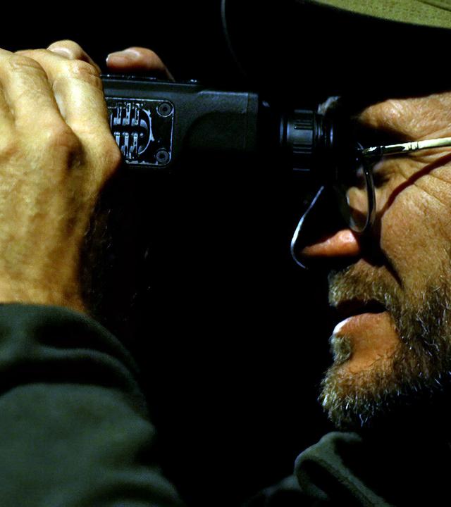 Nahaufnahme eines Mannes im Profil, der nachts durch ein Fernglas schaut. Er trägt eine Brille und eine Mütze, und seine Hand hält das Fernglas fest, während er konzentriert in die Dunkelheit blickt.