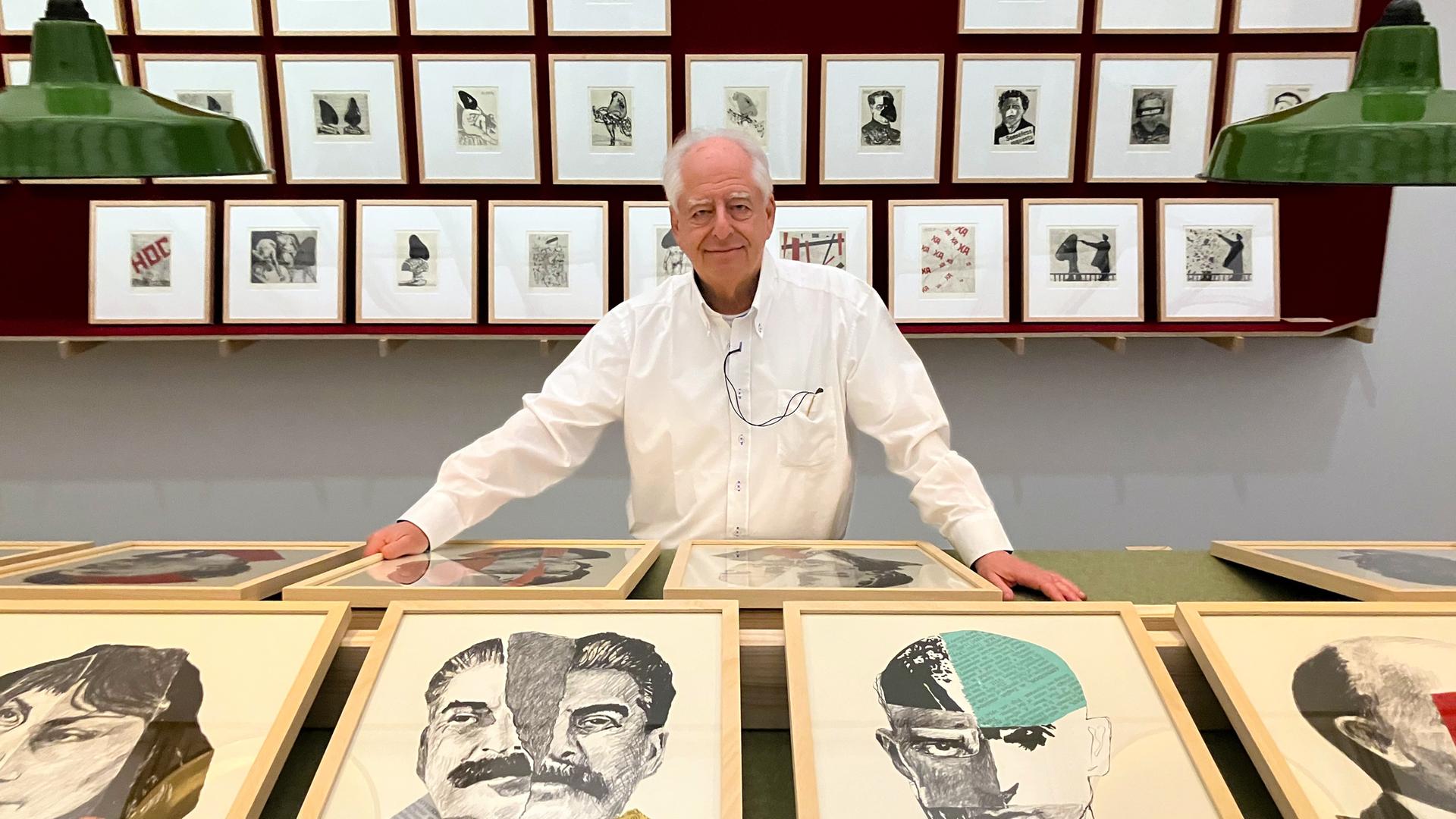 Ein älterer Mann mit weißem Haarkranz und weißem Hemd steht lächelnd hinter einem Podest, auf dem zwei Reihen Bilder präsentiert werden. Hinter ihm an der Wand hängen kleinere gerahmte Bilder in drei Reihen übereinander.