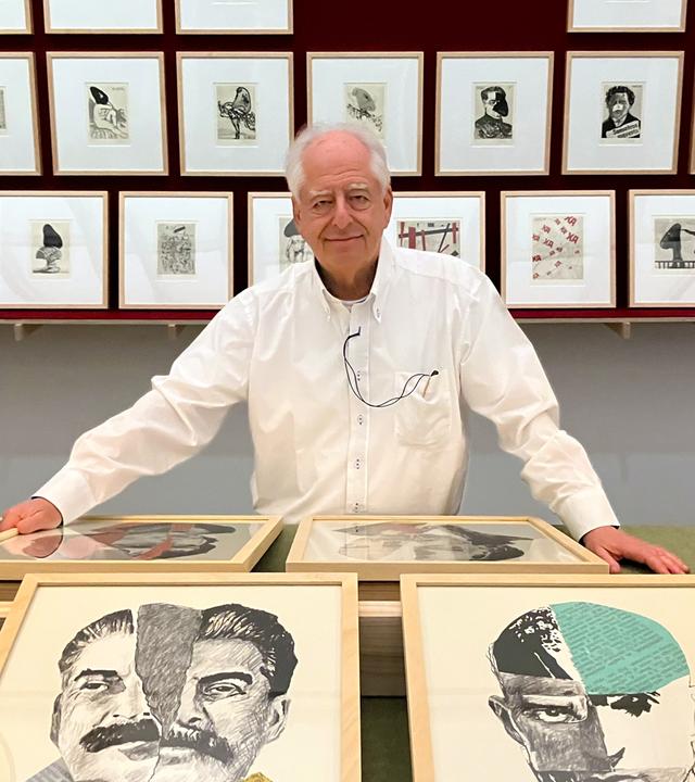 Ein älterer Mann mit weißem Haarkranz und weißem Hemd steht lächelnd hinter einem Podest, auf dem zwei Reihen Bilder präsentiert werden. Hinter ihm an der Wand hängen kleinere gerahmte Bilder in drei Reihen übereinander.