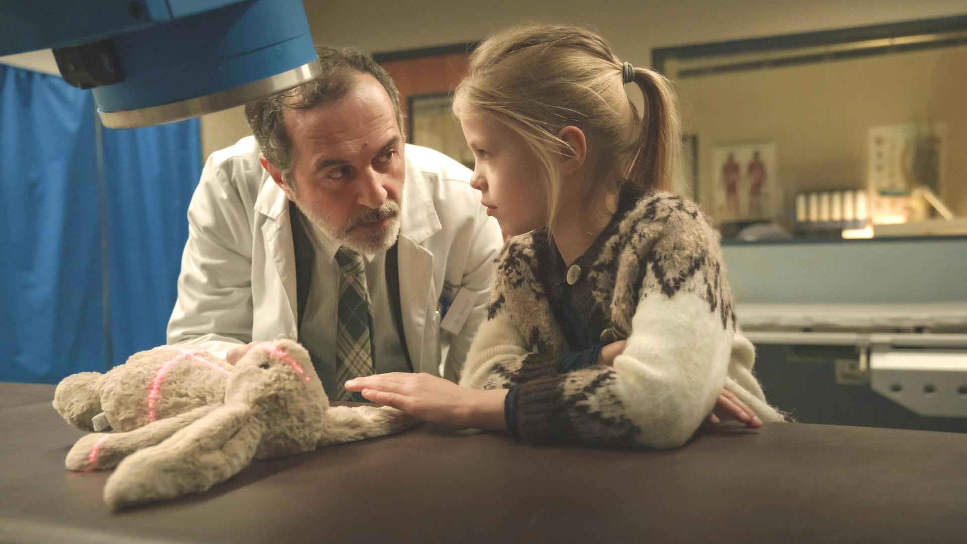 "Doktor Ballouz - Willkommen zurück": Doktor Ballouz (Merab Ninidze) und Flori (Mavie Meschkowski) stehen vor einem Röntgengerät, unter dem ein Stoffhase liegt, auf den rote Lichtstreifen fallen. Der Arzt sieht das Mädchen eindringlich an, das schüchtern seine Hand in Richtung des Hasen legt.