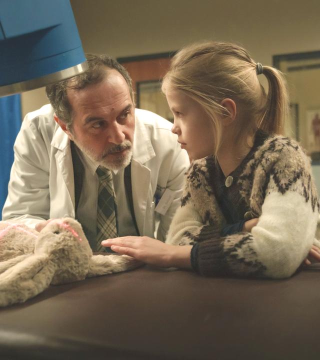 "Doktor Ballouz - Willkommen zurück": Doktor Ballouz (Merab Ninidze) und Flori (Mavie Meschkowski) stehen vor einem Röntgengerät, unter dem ein Stoffhase liegt, auf den rote Lichtstreifen fallen. Der Arzt sieht das Mädchen eindringlich an, das schüchtern seine Hand in Richtung des Hasen legt.