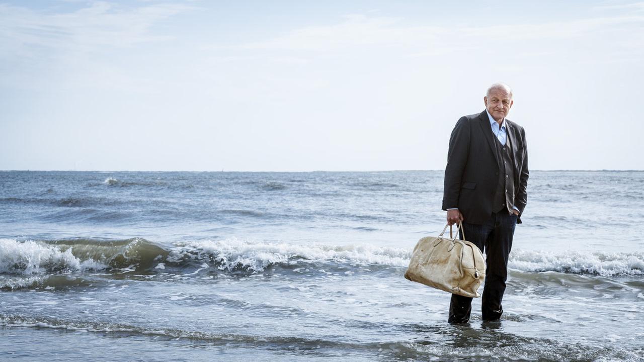 Wilsberg steht im Anzug und mit einer beigen Reisetasche am Strand, seine Füße sind von Wasser umspült.