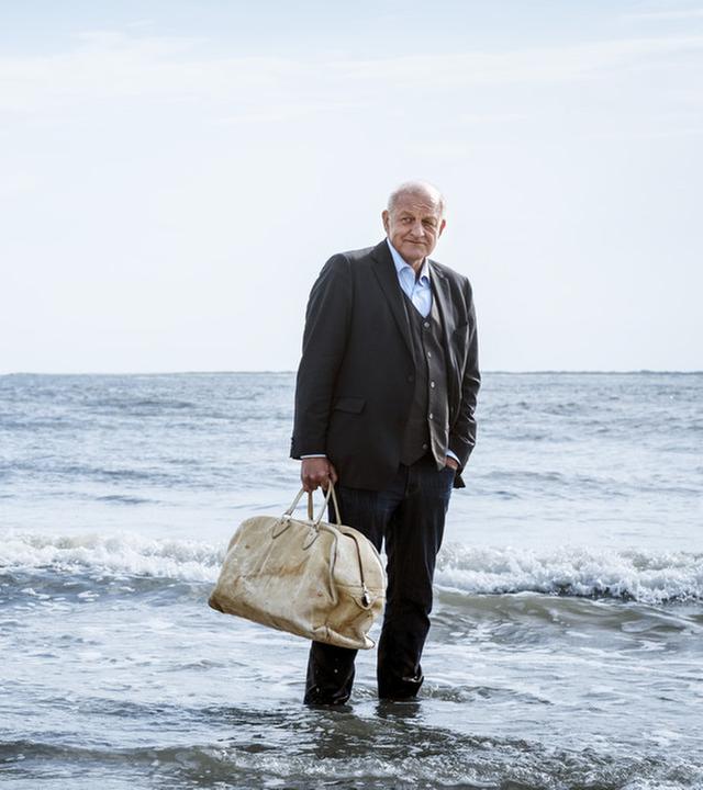 Wilsberg steht im Anzug und mit einer beigen Reisetasche am Strand, seine Füße sind von Wasser umspült.