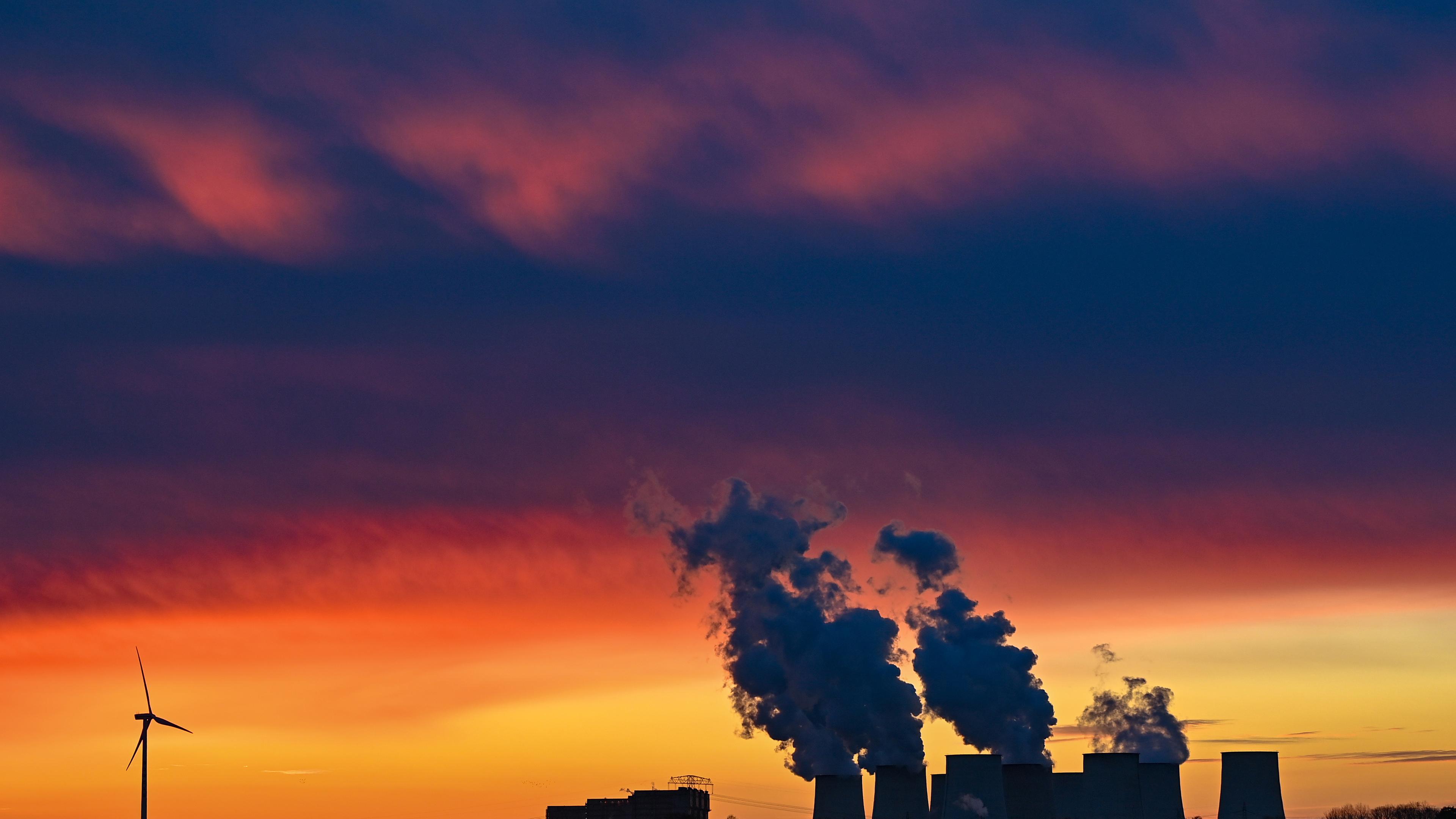 Vor dem Sonnenuntergang heben sich dunkel die Silhouetten eines Braunkohlekraftwerks mit rauchenden Türmen und eines Windrads ab.