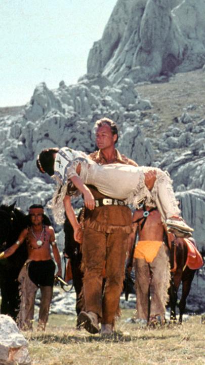 Old Shatterhand (Lex Barker) hält die leblose Nscho-tschi (Marie Versini) in seinem Armen, Winnetou schreitet neben ihm. Im Hintergrund sind mehrere Apachen vor einer Felsformation zu sehen.