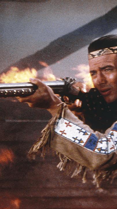 Winnetou (Pierre Brice) steht vor einer brennenden Hütte und schießt mit einem Gewehr.