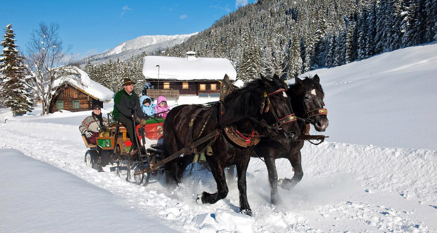 Vor blauem Himmel an dick verschneiten Berghütten vorbei in einem bunten Schlitten, der von zwei Pferden gezogen wird