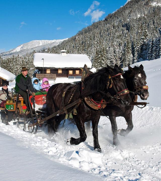 Vor blauem Himmel an dick verschneiten Berghütten vorbei in einem bunten Schlitten, der von zwei Pferden gezogen wird