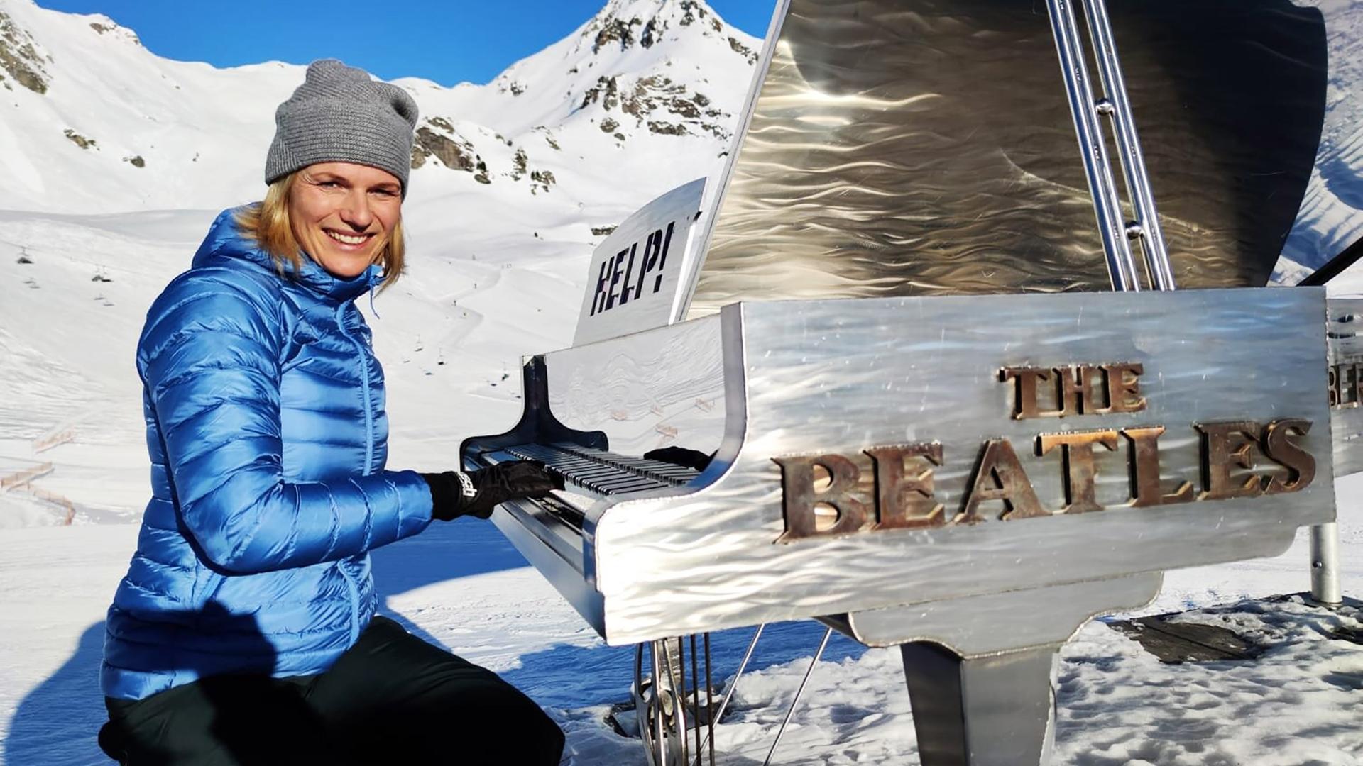 Eine winterlich gekleidete Frau spielt in einer verschneiten Berglandschaft an einem silbernen Flügel