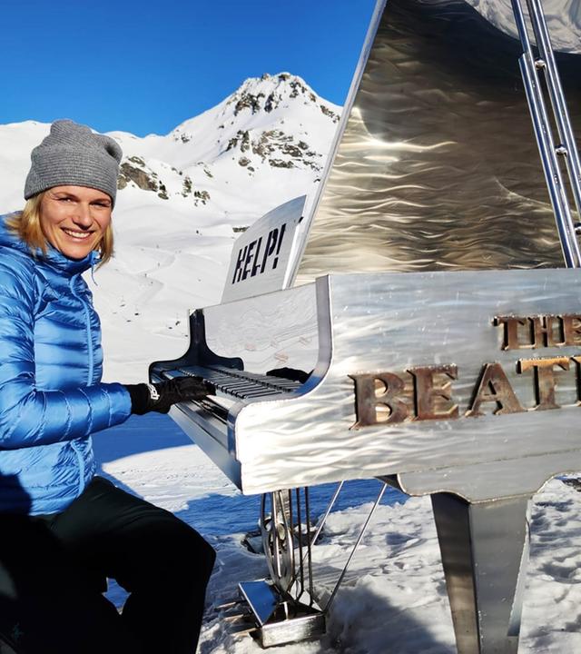 Eine winterlich gekleidete Frau spielt in einer verschneiten Berglandschaft an einem silbernen Flügel